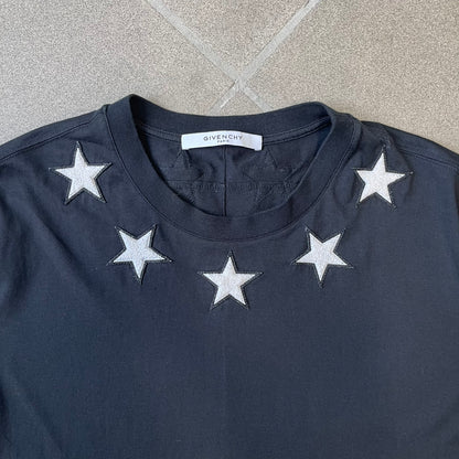Givenchy 2017 Black Star Patch 4-7 Tee is a black, Cuban-fit T-shirt made from 100% cotton, featuring fuzzy white star patches trimming the collar on both the front and back. White "4" and "7" numbering is printed on the sleeves.