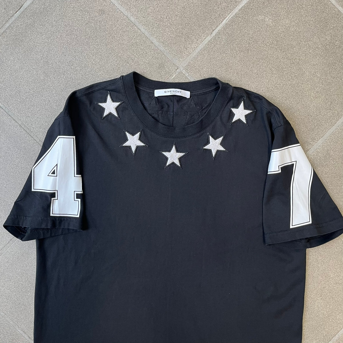 Givenchy 2017 Black Star Patch 4-7 Tee is a black, Cuban-fit T-shirt made from 100% cotton, featuring fuzzy white star patches trimming the collar on both the front and back. White "4" and "7" numbering is printed on the sleeves.