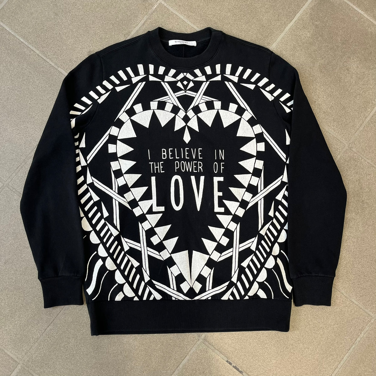 Givenchy FW16 "I Believe in the Power of Love" Crewneck is a black crewneck made from 100% cotton, featuring a white graphic heart built from varying patterns that grow from small to large, with "I Believe in the Power of Love" text placed at its center.&nbsp;