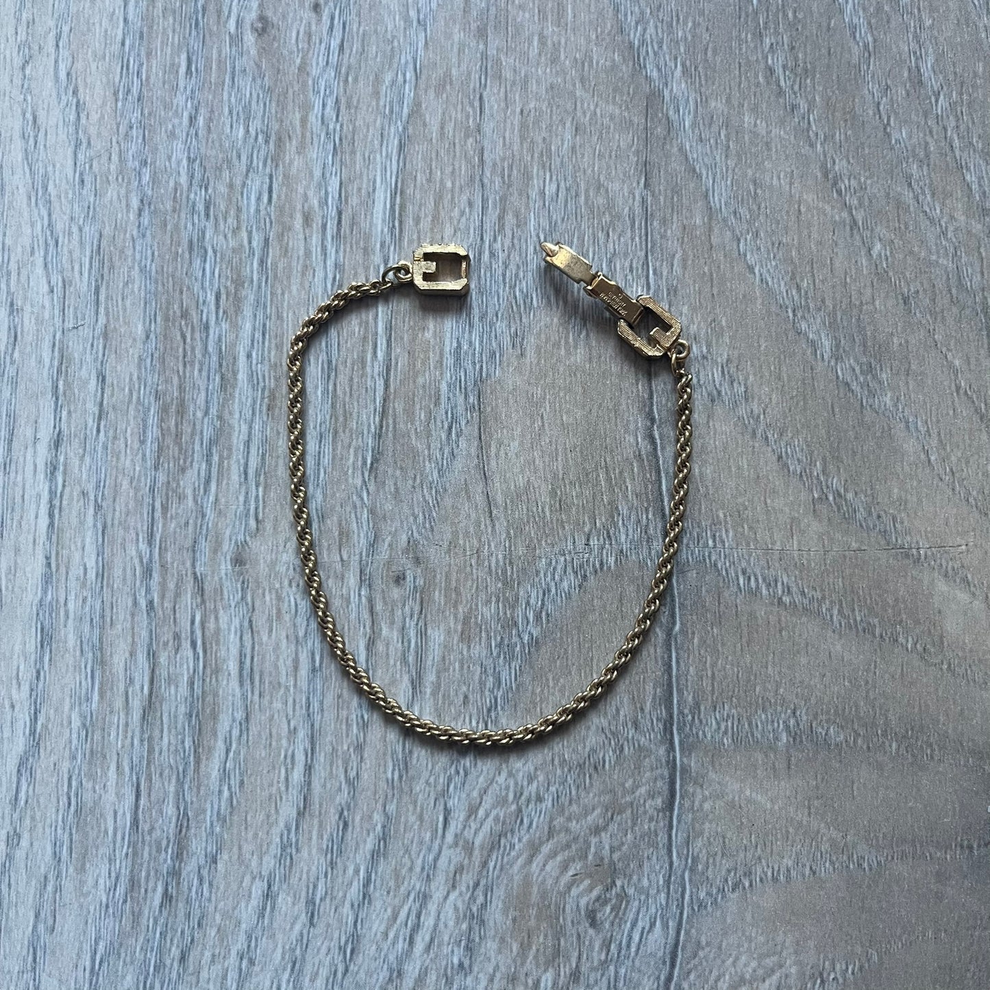 The Givenchy Gold-Tone Vintage Rope Bracelet features a thin gold-tone rope-style chain with larger circular end links shaped like the Givenchy “G” connecting to the clasp. The foldover clasp is oversized compared to the chain and is stamped Givenchy Paris New York. Vintage piece with a uniform gold-tone finish.