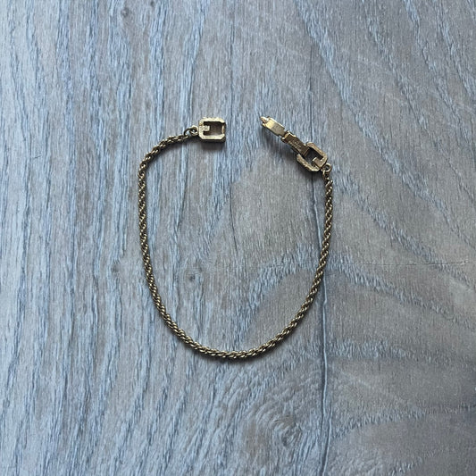 The Givenchy Gold-Tone Vintage Rope Bracelet features a thin gold-tone rope-style chain with larger circular end links shaped like the Givenchy “G” connecting to the clasp. The foldover clasp is oversized compared to the chain and is stamped Givenchy Paris New York. Vintage piece with a uniform gold-tone finish.