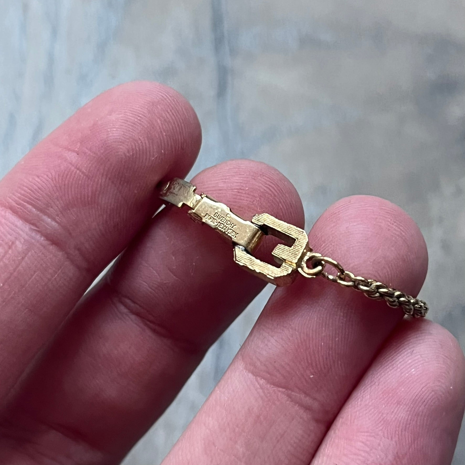The Givenchy Gold-Tone Vintage Rope Bracelet features a thin gold-tone rope-style chain with larger circular end links shaped like the Givenchy “G” connecting to the clasp. The foldover clasp is oversized compared to the chain and is stamped Givenchy Paris New York. Vintage piece with a uniform gold-tone finish.