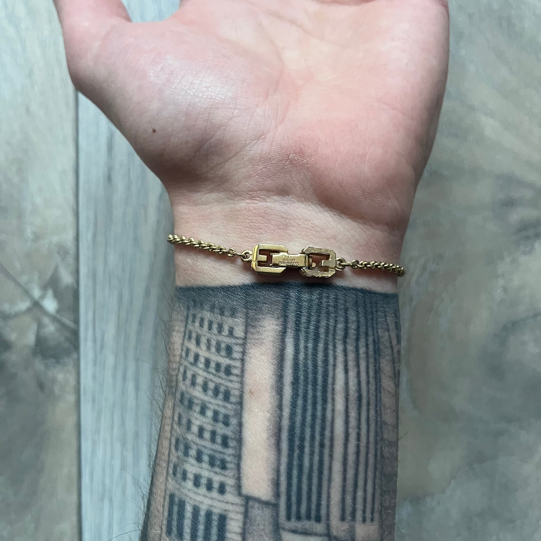 The Givenchy Gold-Tone Vintage Rope Bracelet features a thin gold-tone rope-style chain with larger circular end links shaped like the Givenchy “G” connecting to the clasp. The foldover clasp is oversized compared to the chain and is stamped Givenchy Paris New York. Vintage piece with a uniform gold-tone finish.