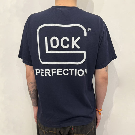 Navy Glock Perfection Tee from the 2000s, made on a Hanes Heavyweight 50/50 cotton-poly blend. Features a large white Glock Perfection logo on the back and a smaller matching logo on the left chest. Thick, high-quality construction.