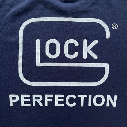 Navy Glock Perfection Tee from the 2000s, made on a Hanes Heavyweight 50/50 cotton-poly blend. Features a large white Glock Perfection logo on the back and a smaller matching logo on the left chest. Thick, high-quality construction.