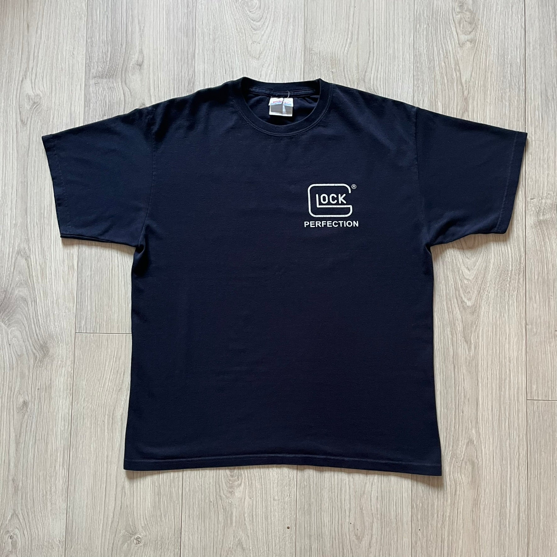 Navy Glock Perfection Tee from the 2000s, made on a Hanes Heavyweight 50/50 cotton-poly blend. Features a large white Glock Perfection logo on the back and a smaller matching logo on the left chest. Thick, high-quality construction.