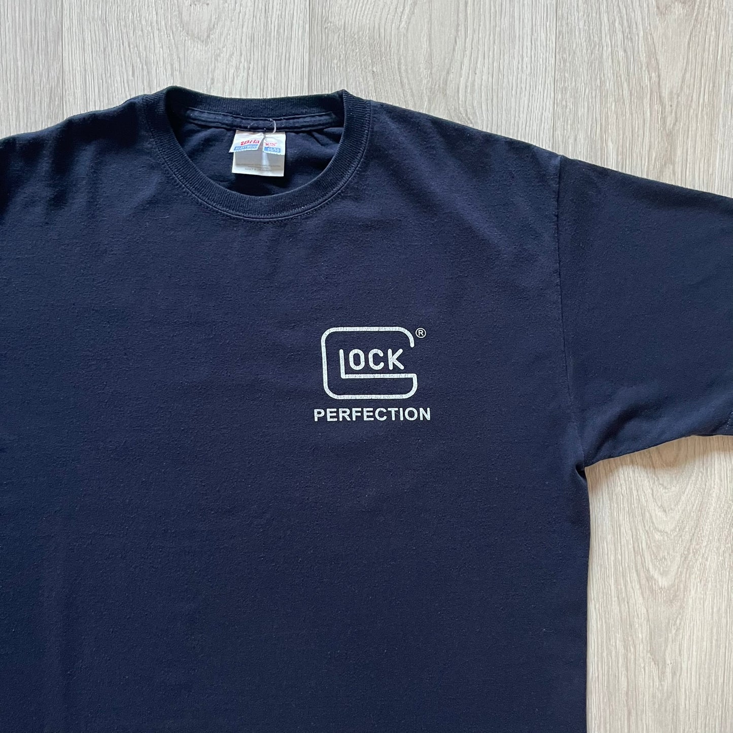 Navy Glock Perfection Tee from the 2000s, made on a Hanes Heavyweight 50/50 cotton-poly blend. Features a large white Glock Perfection logo on the back and a smaller matching logo on the left chest. Thick, high-quality construction.