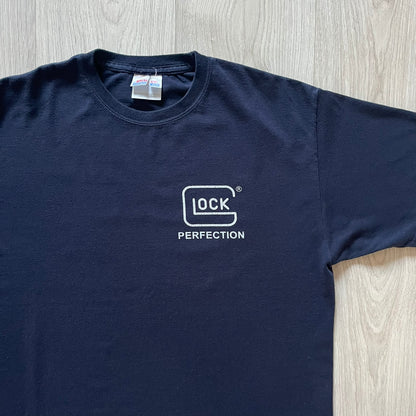 Navy Glock Perfection Tee from the 2000s, made on a Hanes Heavyweight 50/50 cotton-poly blend. Features a large white Glock Perfection logo on the back and a smaller matching logo on the left chest. Thick, high-quality construction.