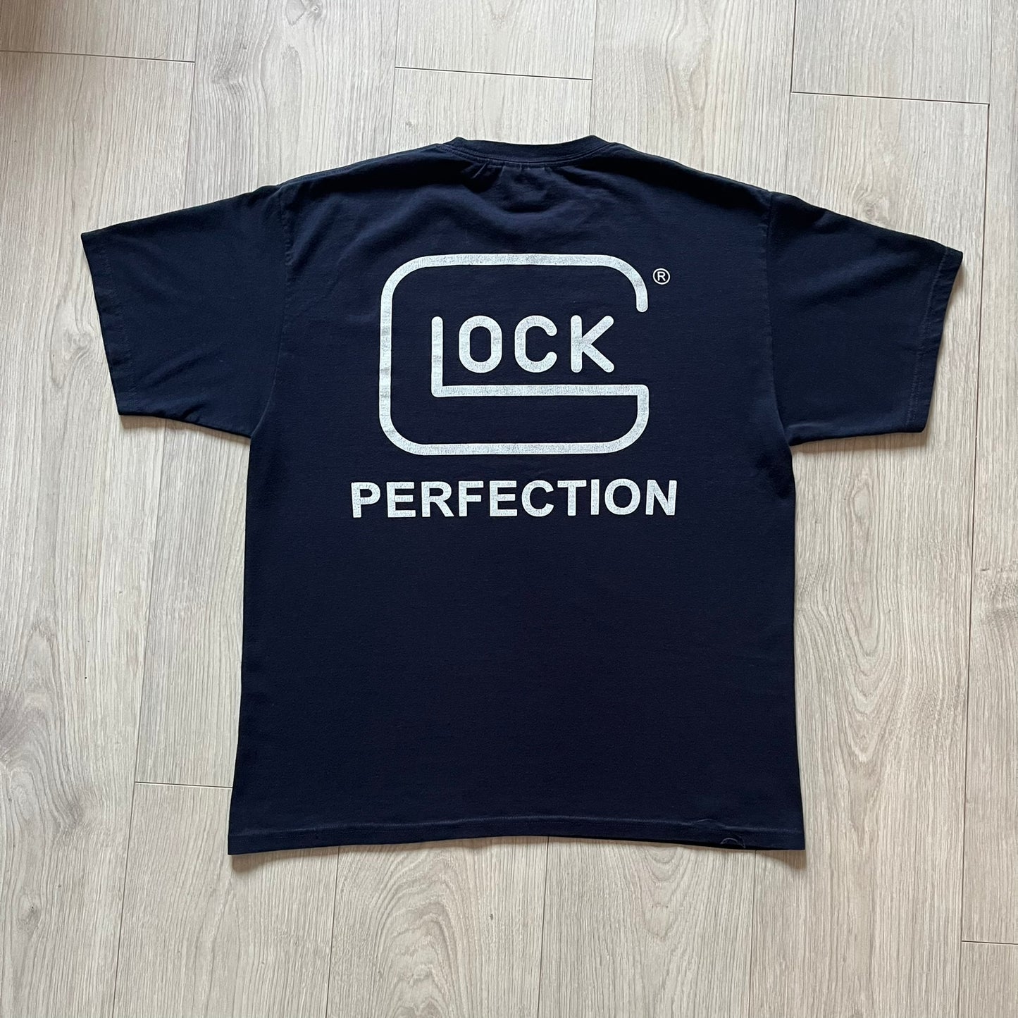 Navy Glock Perfection Tee from the 2000s, made on a Hanes Heavyweight 50/50 cotton-poly blend. Features a large white Glock Perfection logo on the back and a smaller matching logo on the left chest. Thick, high-quality construction.
