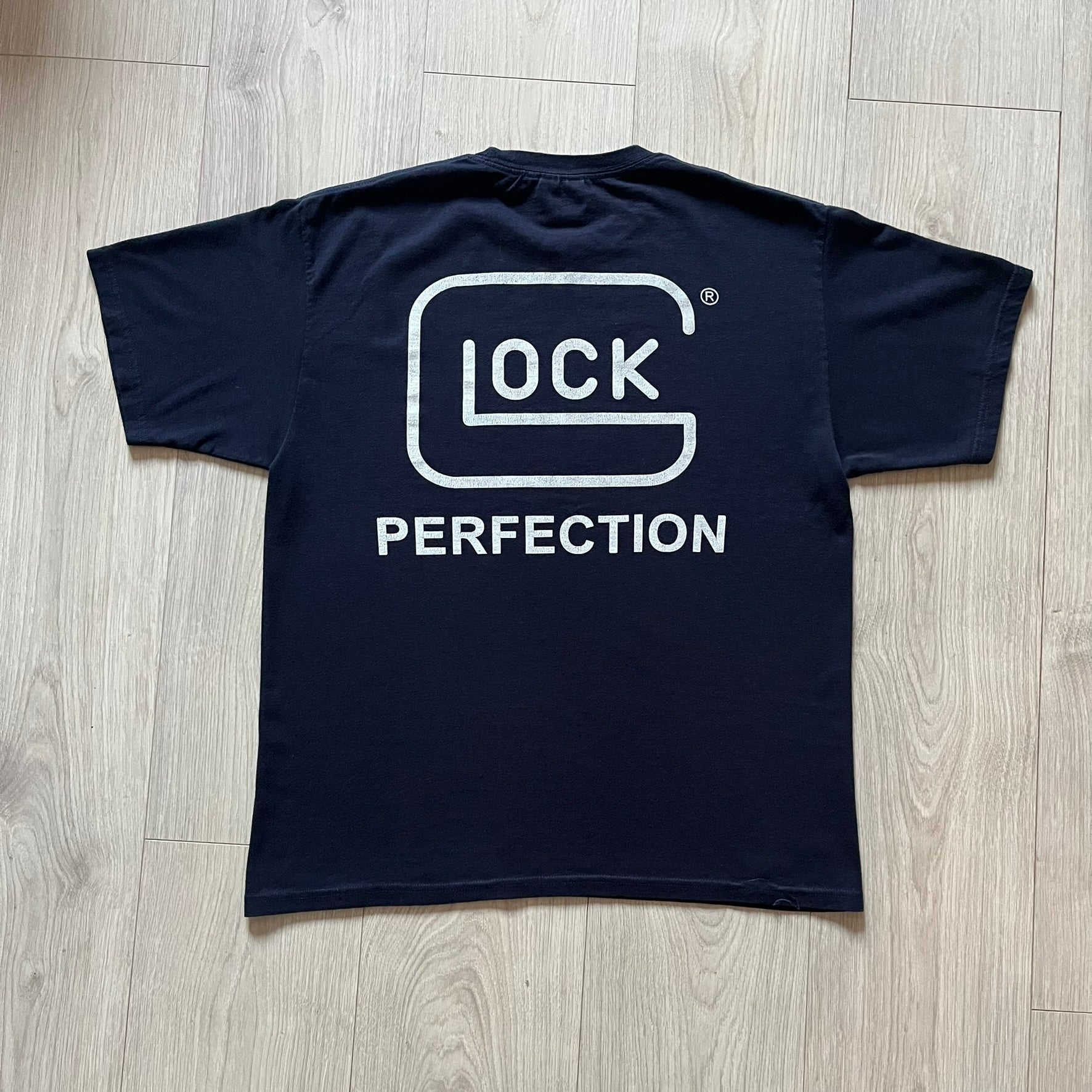 Navy Glock Perfection Tee from the 2000s, made on a Hanes Heavyweight 50/50 cotton-poly blend. Features a large white Glock Perfection logo on the back and a smaller matching logo on the left chest. Thick, high-quality construction.