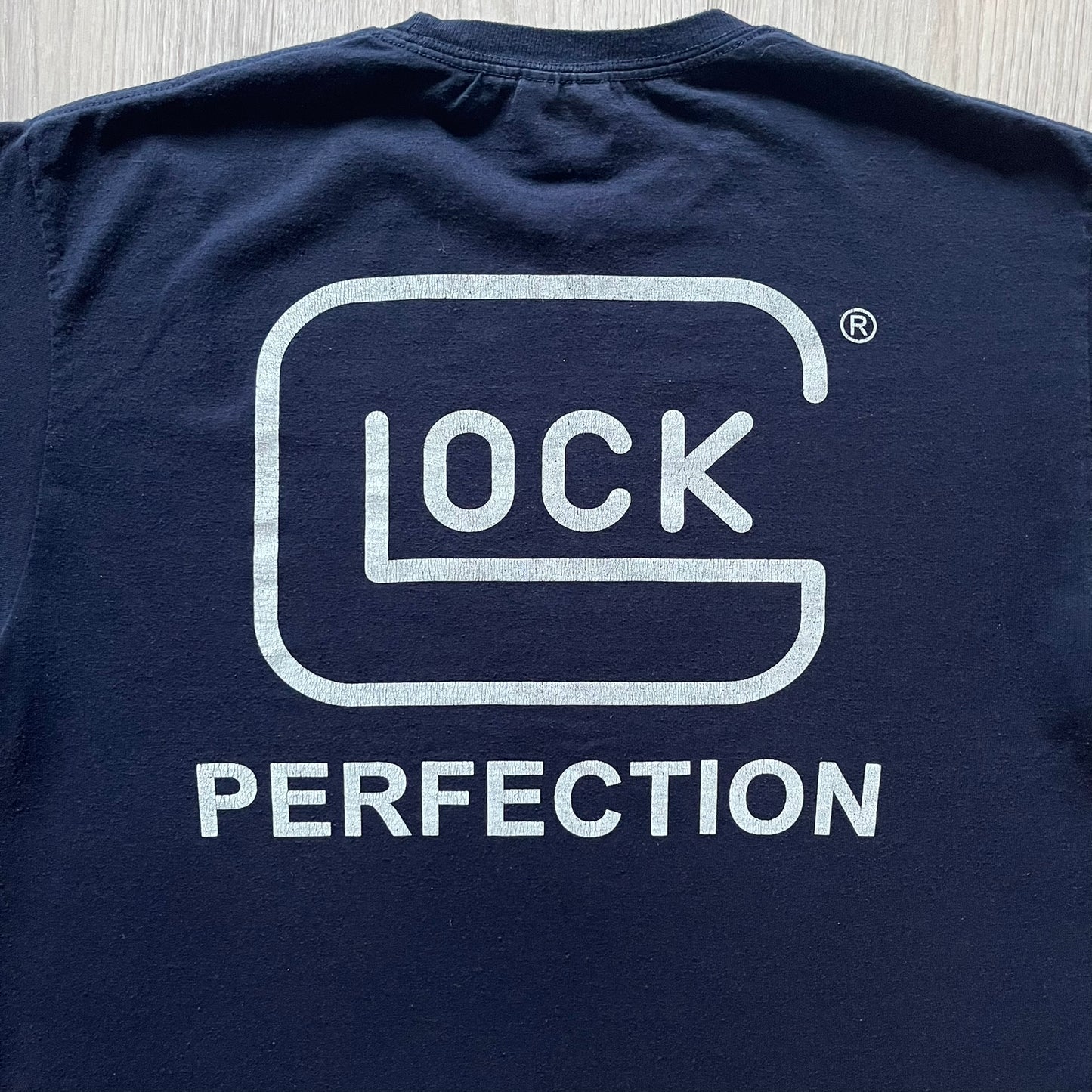 Navy Glock Perfection Tee from the 2000s, made on a Hanes Heavyweight 50/50 cotton-poly blend. Features a large white Glock Perfection logo on the back and a smaller matching logo on the left chest. Thick, high-quality construction.