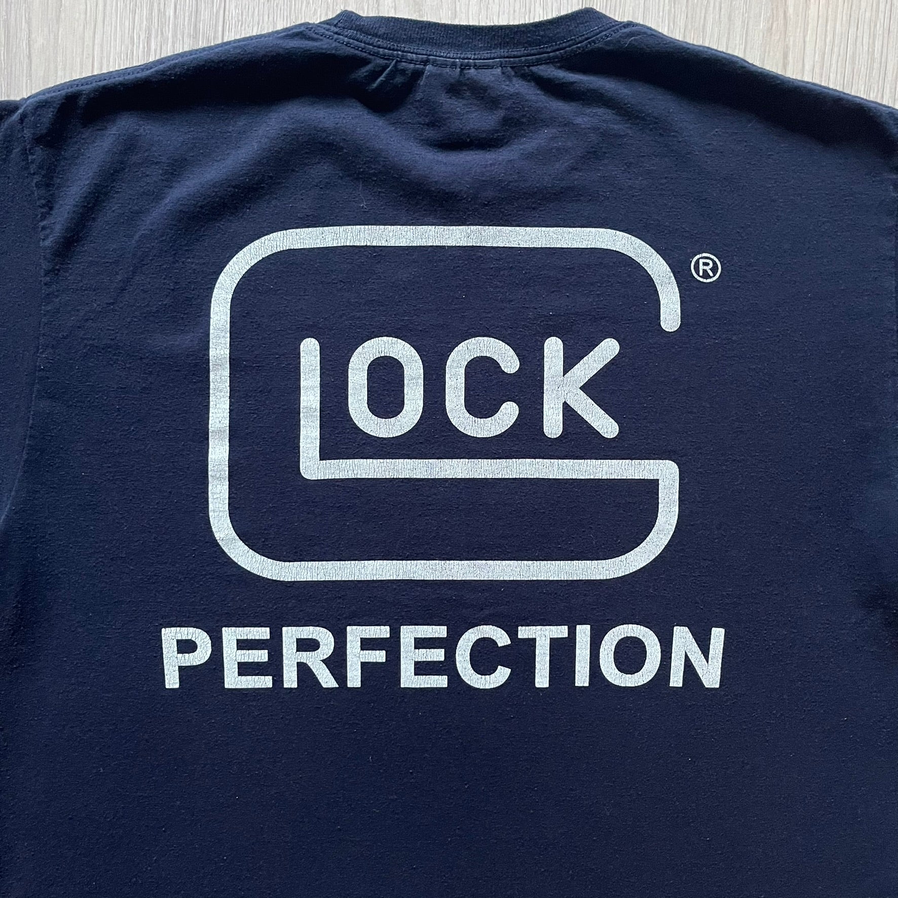 Navy Glock Perfection Tee from the 2000s, made on a Hanes Heavyweight 50/50 cotton-poly blend. Features a large white Glock Perfection logo on the back and a smaller matching logo on the left chest. Thick, high-quality construction.
