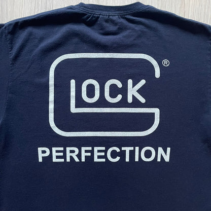 Navy Glock Perfection Tee from the 2000s, made on a Hanes Heavyweight 50/50 cotton-poly blend. Features a large white Glock Perfection logo on the back and a smaller matching logo on the left chest. Thick, high-quality construction.
