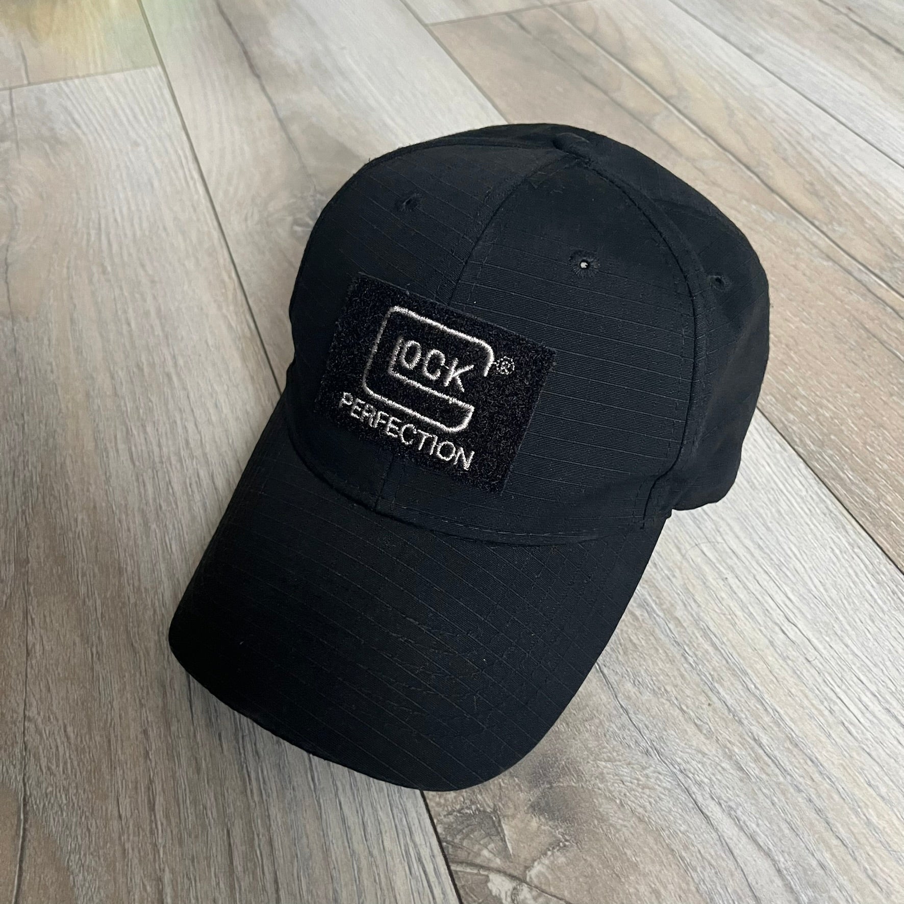 Glock Perfection USA Flag Baseball Cap is a black baseball cap featuring a fuzzy-style front patch with a white Glock Perfection logo. The left side has a silver USA flag patch, and the back is finished with an additional Glock Perfection logo. The cap has a classic curved brim and standard adjustable fit.