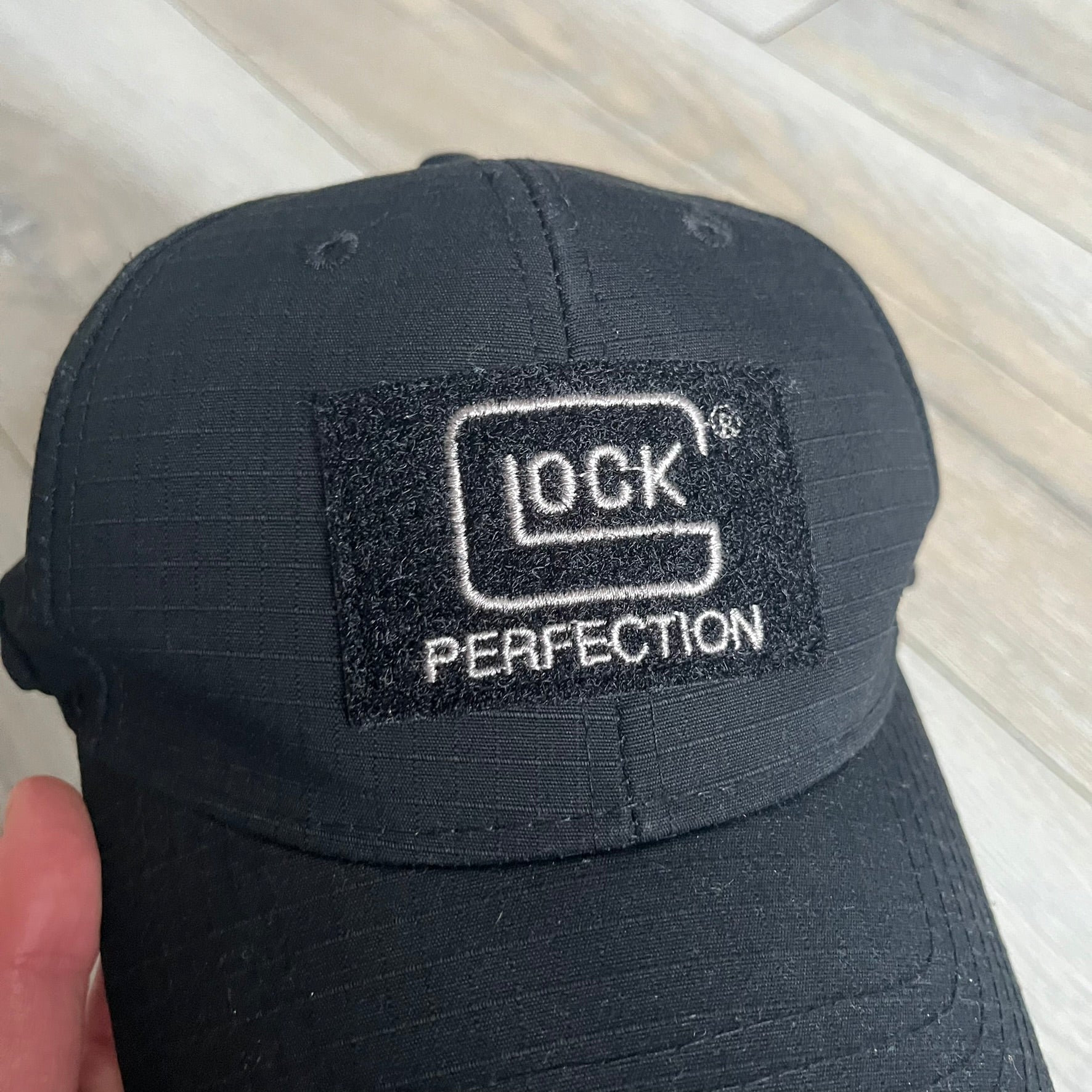 Glock Perfection USA Flag Baseball Cap is a black baseball cap featuring a fuzzy-style front patch with a white Glock Perfection logo. The left side has a silver USA flag patch, and the back is finished with an additional Glock Perfection logo. The cap has a classic curved brim and standard adjustable fit.