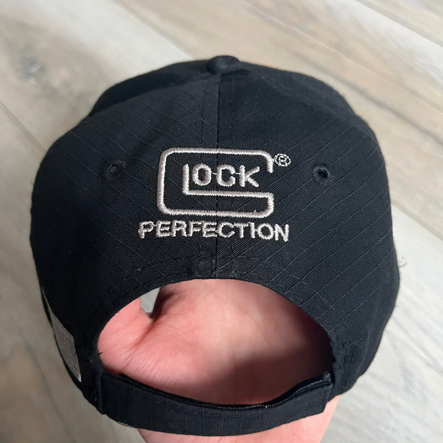 Glock Perfection USA Flag Baseball Cap is a black baseball cap featuring a fuzzy-style front patch with a white Glock Perfection logo. The left side has a silver USA flag patch, and the back is finished with an additional Glock Perfection logo. The cap has a classic curved brim and standard adjustable fit.