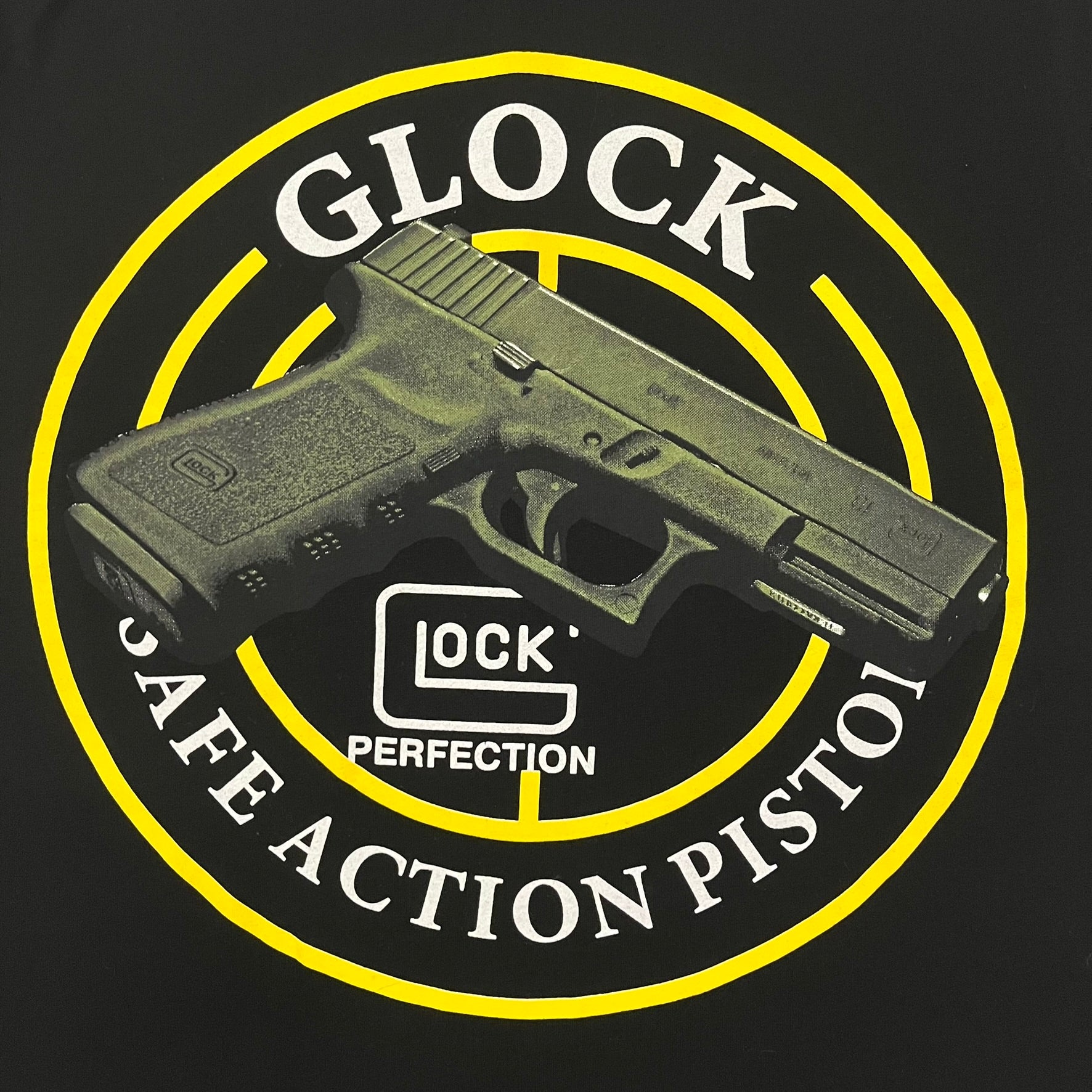 Glock Safe Action Pistol T-Shirt in black, made on an Orijin blank from 100% cotton. Features a large Glock pistol graphic on the front set inside a yellow circle, with “Glock Safe Action” text and Glock logo running around the graphic. The back has a white Glock logo printed below the collar.&nbsp;