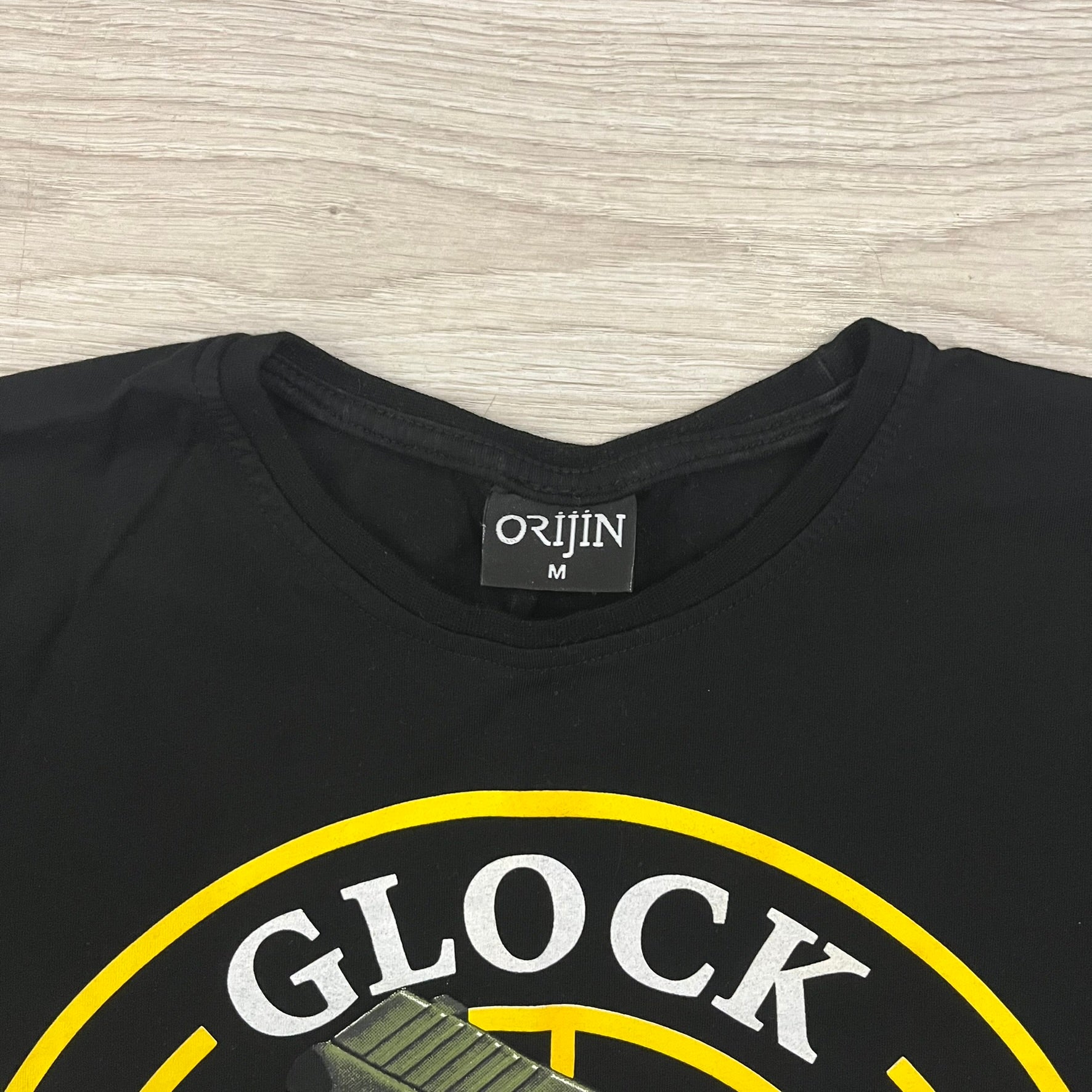 Glock Safe Action Pistol T-Shirt in black, made on an Orijin blank from 100% cotton. Features a large Glock pistol graphic on the front set inside a yellow circle, with “Glock Safe Action” text and Glock logo running around the graphic. The back has a white Glock logo printed below the collar.&nbsp;
