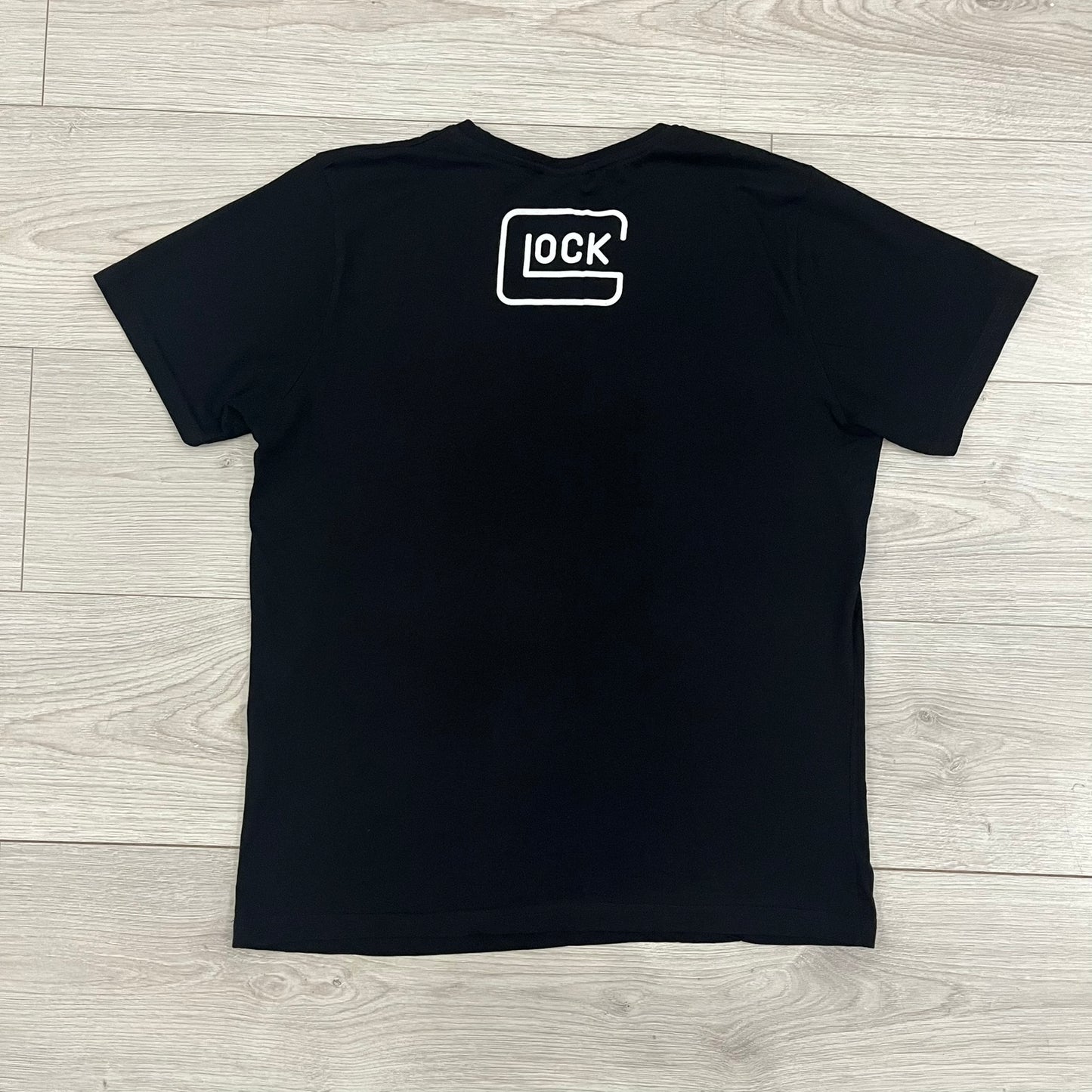 Glock Safe Action Pistol T-Shirt in black, made on an Orijin blank from 100% cotton. Features a large Glock pistol graphic on the front set inside a yellow circle, with “Glock Safe Action” text and Glock logo running around the graphic. The back has a white Glock logo printed below the collar.&nbsp;