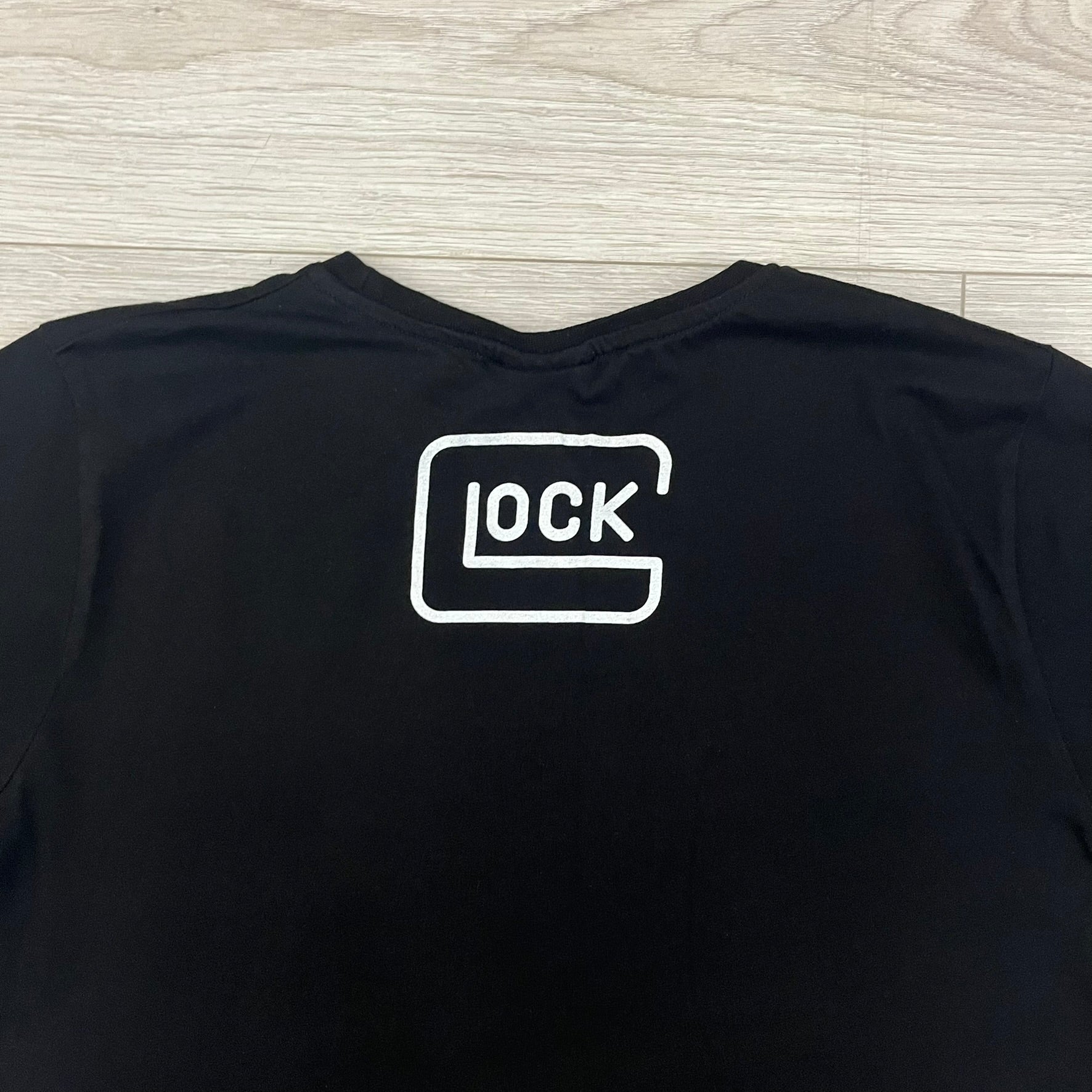 Glock Safe Action Pistol T-Shirt in black, made on an Orijin blank from 100% cotton. Features a large Glock pistol graphic on the front set inside a yellow circle, with “Glock Safe Action” text and Glock logo running around the graphic. The back has a white Glock logo printed below the collar.&nbsp;