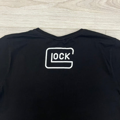 Glock Safe Action Pistol T-Shirt in black, made on an Orijin blank from 100% cotton. Features a large Glock pistol graphic on the front set inside a yellow circle, with “Glock Safe Action” text and Glock logo running around the graphic. The back has a white Glock logo printed below the collar.&nbsp;