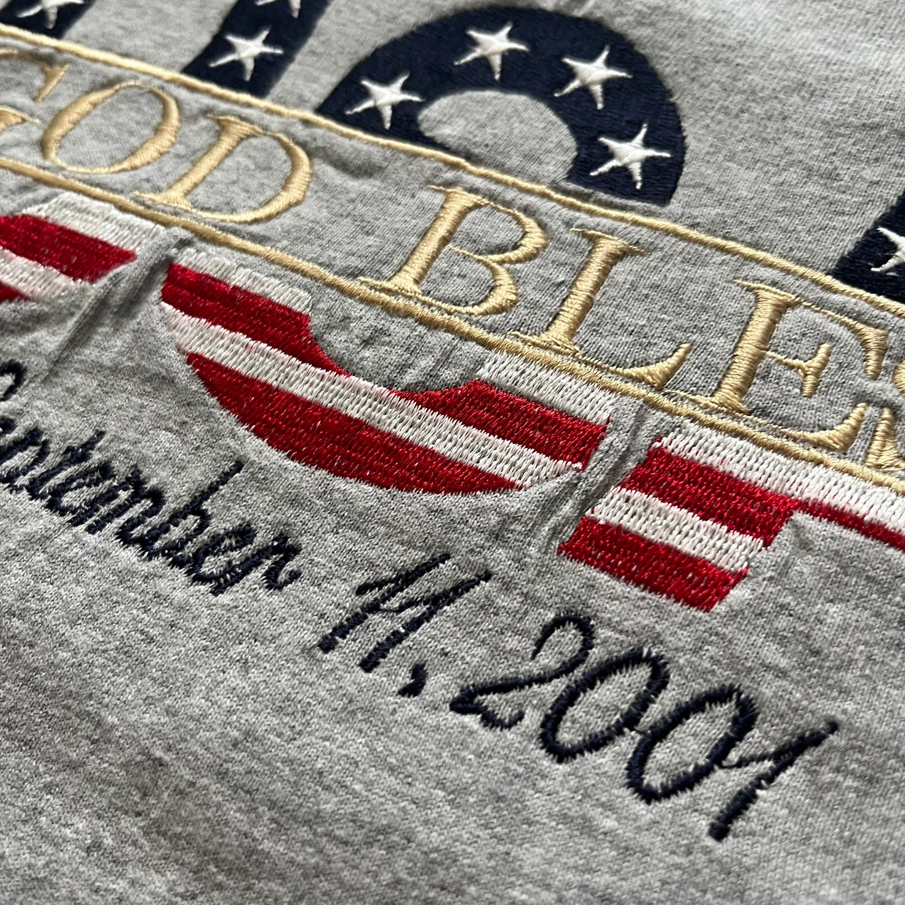 Gray “God Bless USA” tee on an Alstyle blank, made from soft, durable cotton. Embroidery showcases USA in flag colors with gold “God Bless” text and a subtle September 11, 2001 detail underneath. A respectful and collectible commemorative shirt.