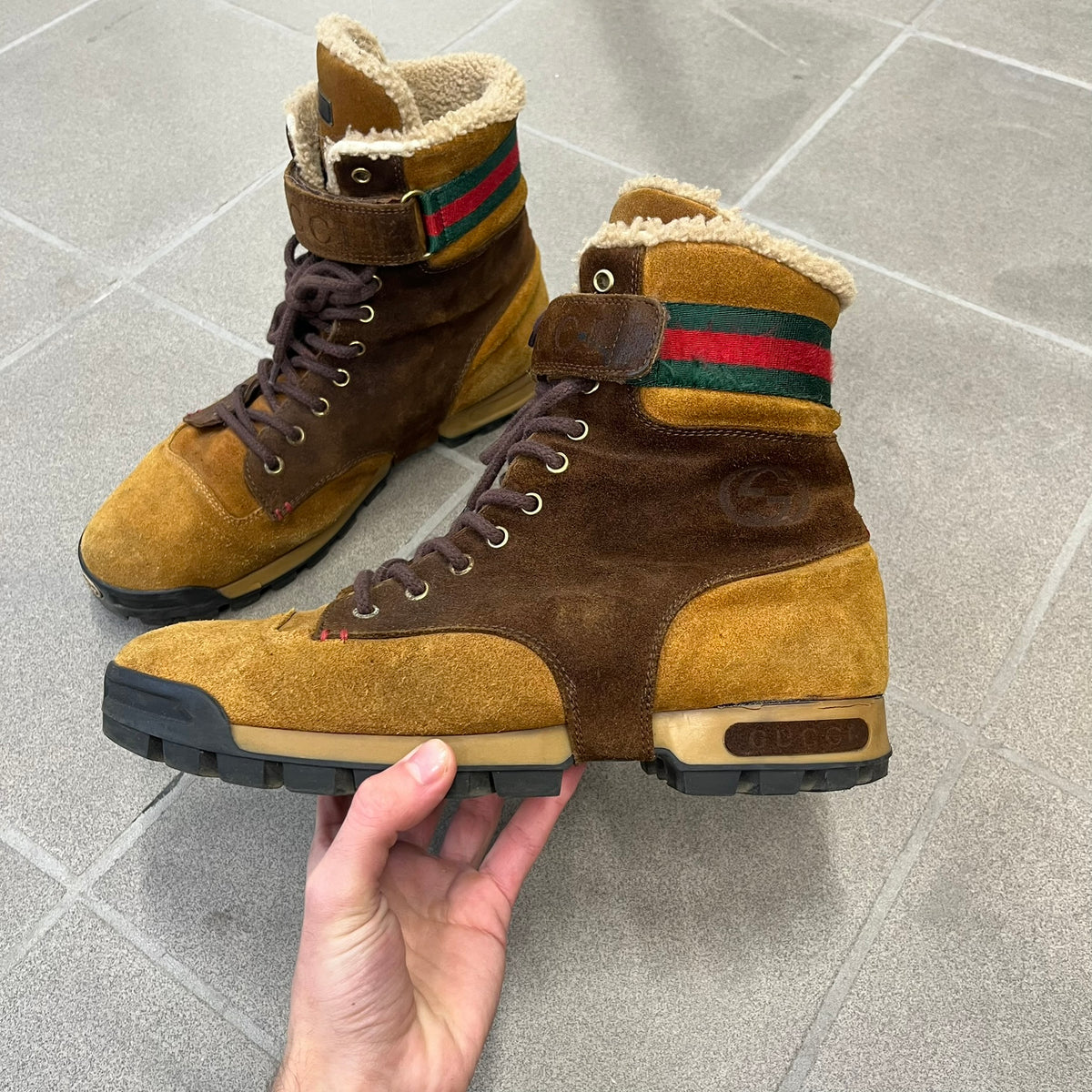 Gucci 2000’s Beige & Brown Shearling Lined Boots are made in Italy from beige and brown suede with a shearling-lined interior. A velcro strap with a Gucci logo closes the top, and the tongue has a leather tab with the Gucci logo. A green and red stripe runs around the top, a GG logo is on the side, and another GG logo appears on the sole along with a small one at the toe tip and a brown leather Gucci logo section on the sole. Due to age, the sole is falling apart and needs either gluing or a sole replacemen