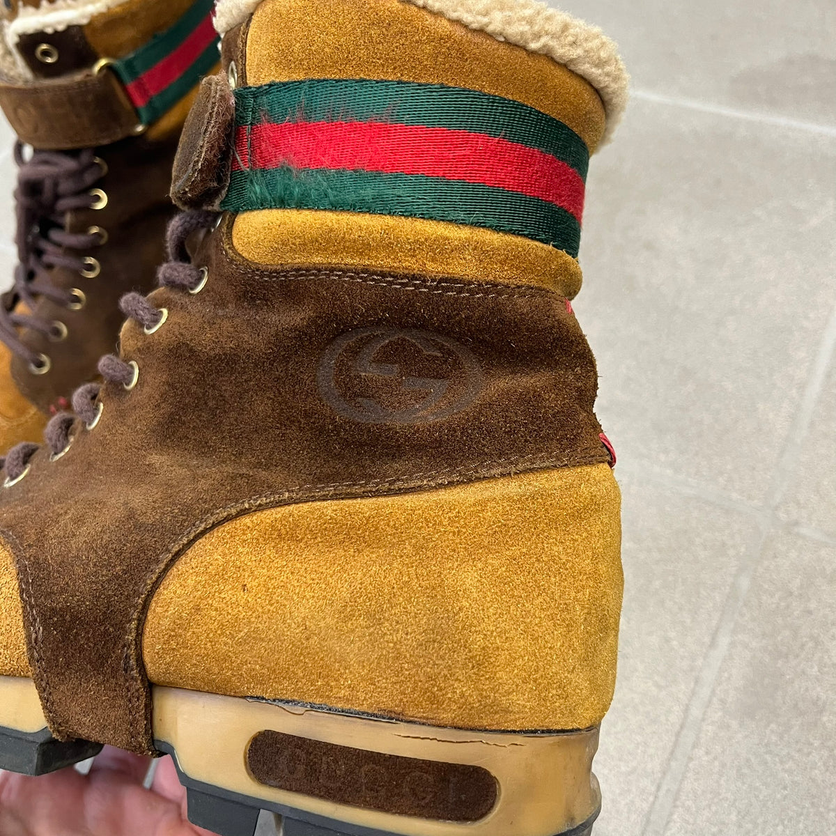 Gucci 2000’s Beige & Brown Shearling Lined Boots are made in Italy from beige and brown suede with a shearling-lined interior. A velcro strap with a Gucci logo closes the top, and the tongue has a leather tab with the Gucci logo. A green and red stripe runs around the top, a GG logo is on the side, and another GG logo appears on the sole along with a small one at the toe tip and a brown leather Gucci logo section on the sole. Due to age, the sole is falling apart and needs either gluing or a sole replacemen