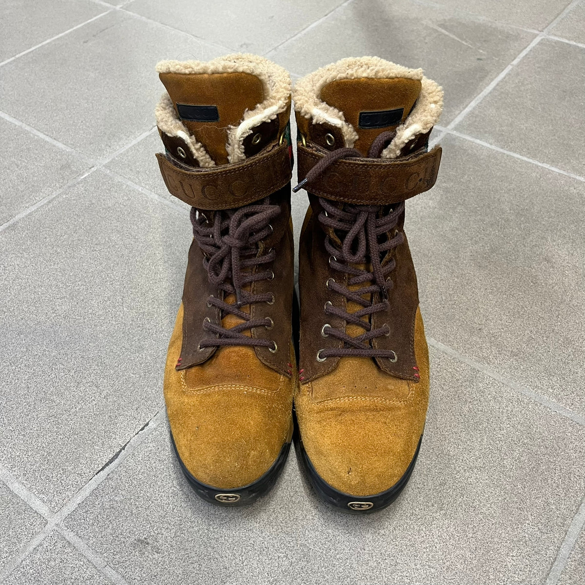 Gucci 2000’s Beige & Brown Shearling Lined Boots are made in Italy from beige and brown suede with a shearling-lined interior. A velcro strap with a Gucci logo closes the top, and the tongue has a leather tab with the Gucci logo. A green and red stripe runs around the top, a GG logo is on the side, and another GG logo appears on the sole along with a small one at the toe tip and a brown leather Gucci logo section on the sole. Due to age, the sole is falling apart and needs either gluing or a sole replacemen