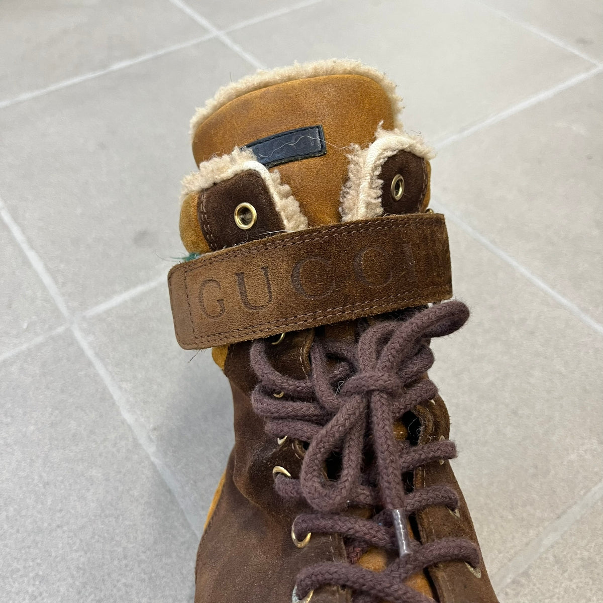 Gucci 2000’s Beige & Brown Shearling Lined Boots are made in Italy from beige and brown suede with a shearling-lined interior. A velcro strap with a Gucci logo closes the top, and the tongue has a leather tab with the Gucci logo. A green and red stripe runs around the top, a GG logo is on the side, and another GG logo appears on the sole along with a small one at the toe tip and a brown leather Gucci logo section on the sole. Due to age, the sole is falling apart and needs either gluing or a sole replacemen