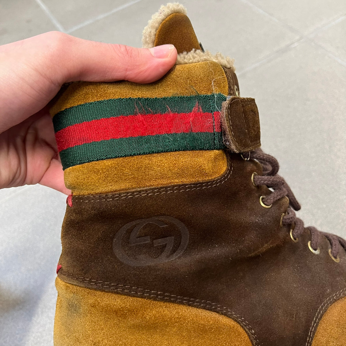 Gucci 2000’s Beige & Brown Shearling Lined Boots are made in Italy from beige and brown suede with a shearling-lined interior. A velcro strap with a Gucci logo closes the top, and the tongue has a leather tab with the Gucci logo. A green and red stripe runs around the top, a GG logo is on the side, and another GG logo appears on the sole along with a small one at the toe tip and a brown leather Gucci logo section on the sole. Due to age, the sole is falling apart and needs either gluing or a sole replacemen