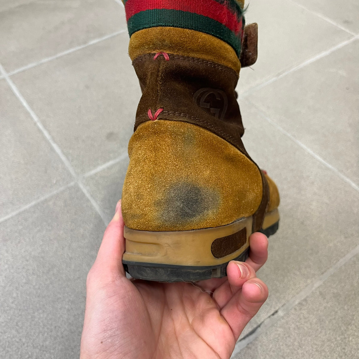 Gucci 2000’s Beige & Brown Shearling Lined Boots are made in Italy from beige and brown suede with a shearling-lined interior. A velcro strap with a Gucci logo closes the top, and the tongue has a leather tab with the Gucci logo. A green and red stripe runs around the top, a GG logo is on the side, and another GG logo appears on the sole along with a small one at the toe tip and a brown leather Gucci logo section on the sole. Due to age, the sole is falling apart and needs either gluing or a sole replacemen