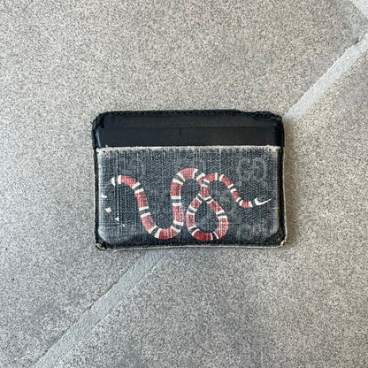 Gucci GG Supreme Kingsnake Card Holder features black and grey GG Supreme canvas with a red and black Kingsnake graphic and black leather trim. It has two card slots on the front, two on the back, and one central slot at the top.
