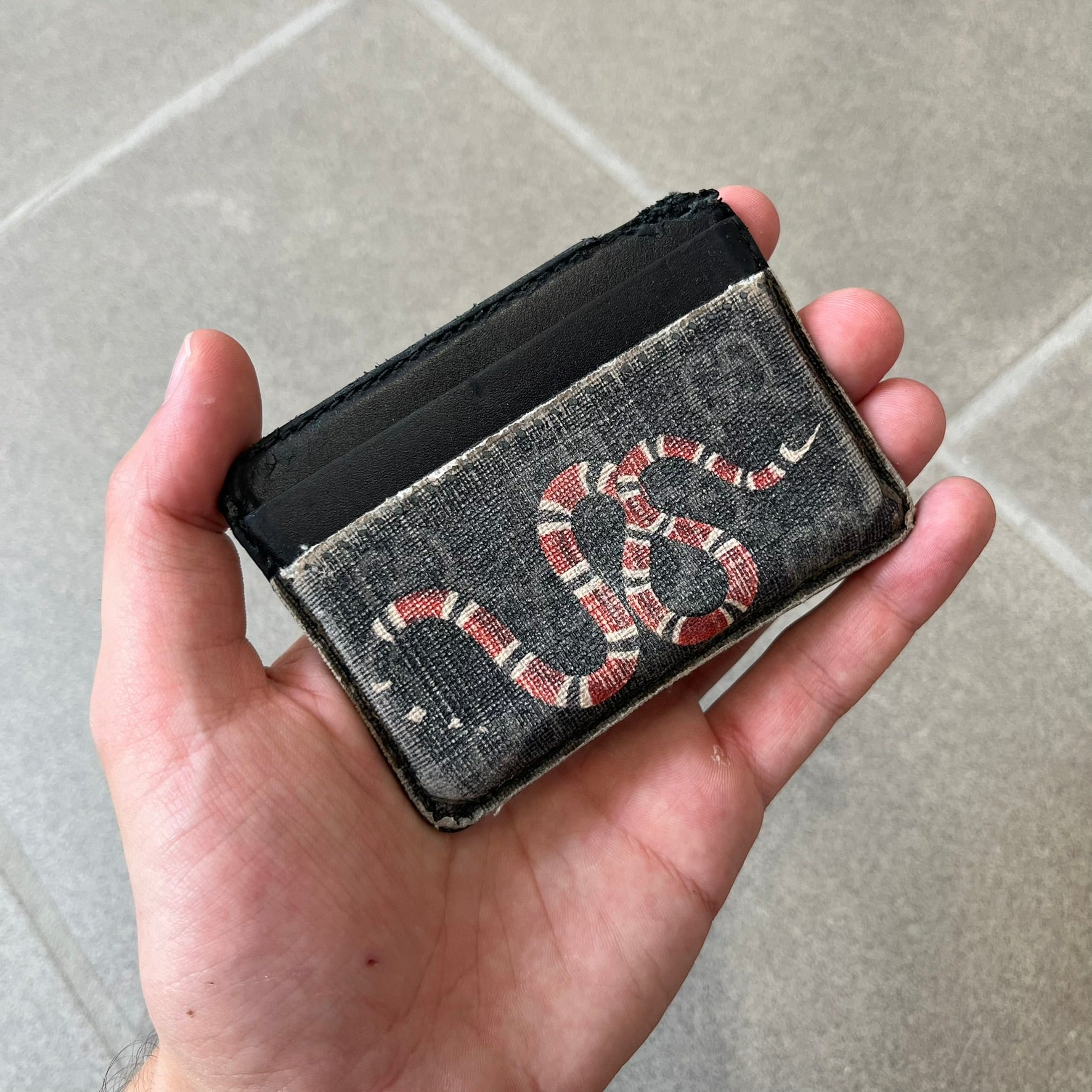 Gucci GG Supreme Kingsnake Card Holder features black and grey GG Supreme canvas with a red and black Kingsnake graphic and black leather trim. It has two card slots on the front, two on the back, and one central slot at the top.
