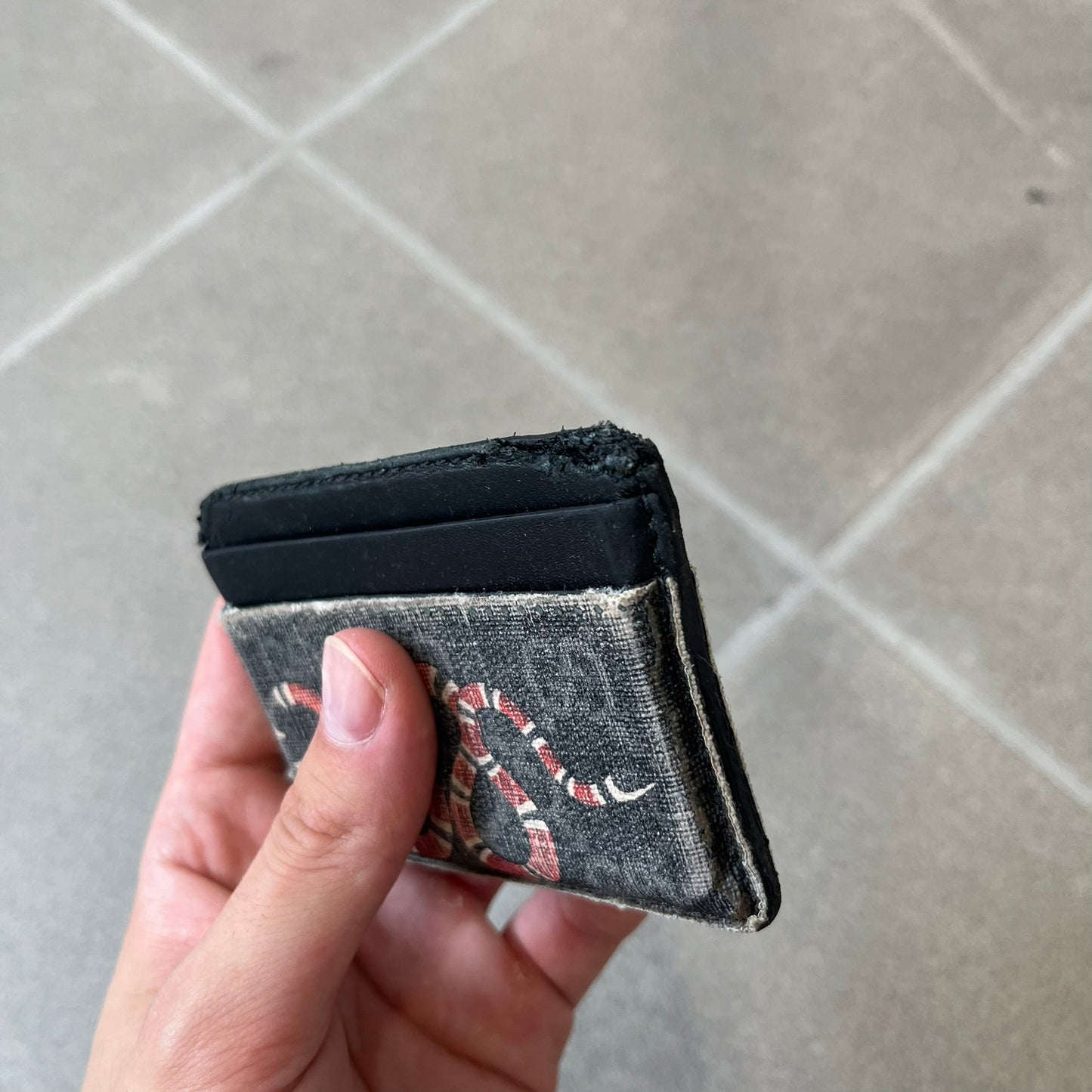 Gucci GG Supreme Kingsnake Card Holder features black and grey GG Supreme canvas with a red and black Kingsnake graphic and black leather trim. It has two card slots on the front, two on the back, and one central slot at the top.
