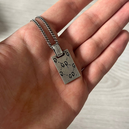 Hand holding a silver necklace with a tag against a neutral background