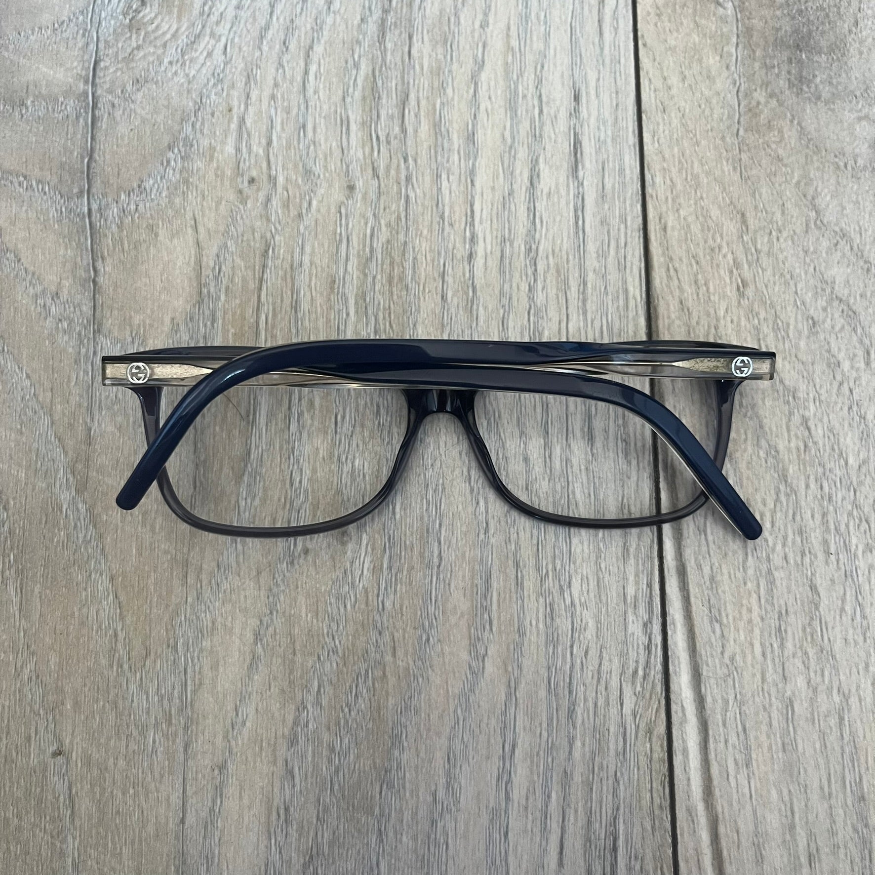 Gucci Gray-Blue Glasses Frames feature a translucent gray and dark blue acetate construction with a clear finish. Small silver GG logos are positioned on the front sides of the frames near the hinges. The frames are sold without lenses and come with a Gucci monogram case and a brown cleaning cloth.