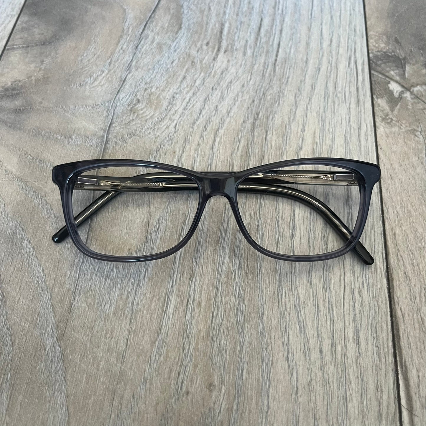 Gucci Gray-Blue Glasses Frames feature a translucent gray and dark blue acetate construction with a clear finish. Small silver GG logos are positioned on the front sides of the frames near the hinges. The frames are sold without lenses and come with a Gucci monogram case and a brown cleaning cloth.