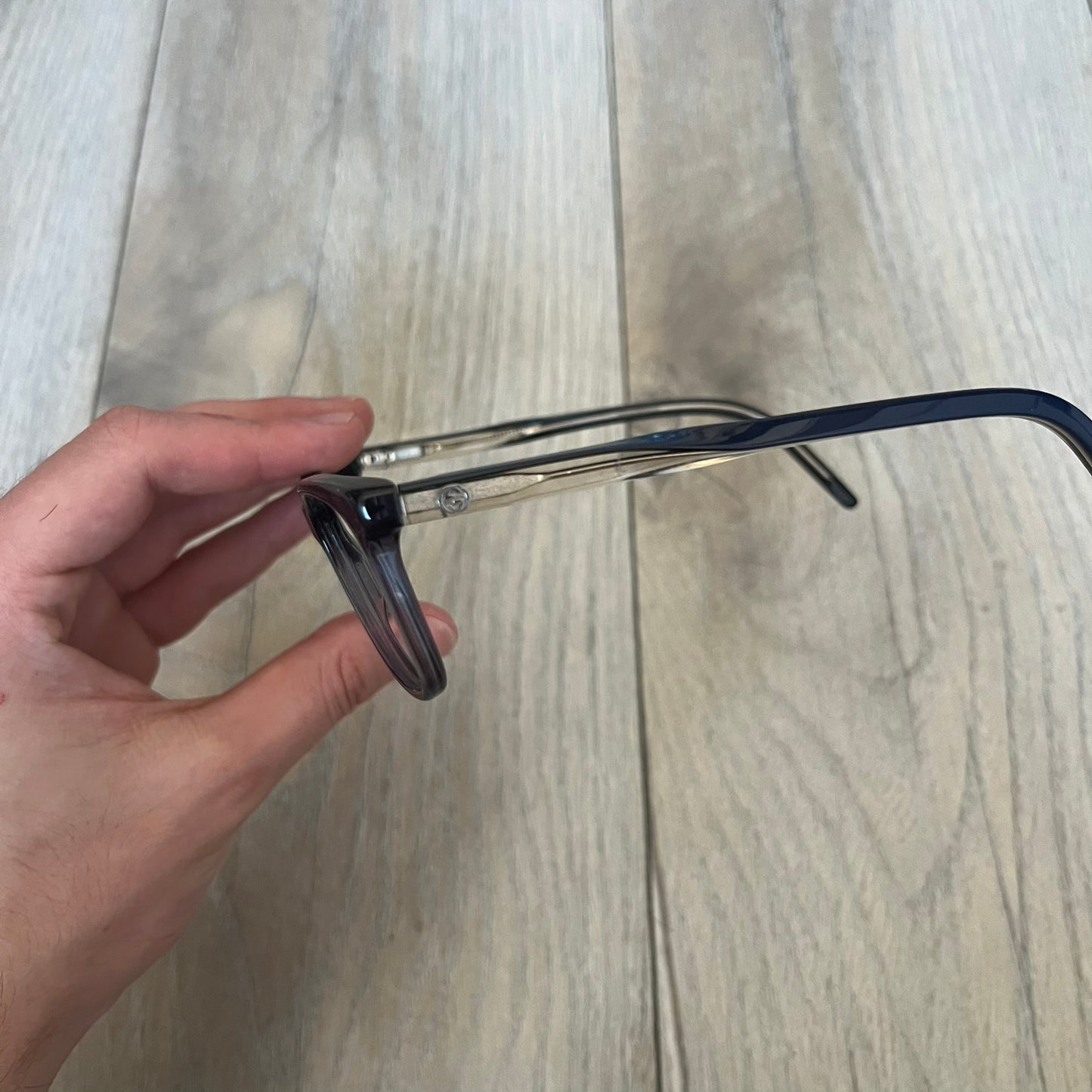 Gucci Gray-Blue Glasses Frames feature a translucent gray and dark blue acetate construction with a clear finish. Small silver GG logos are positioned on the front sides of the frames near the hinges. The frames are sold without lenses and come with a Gucci monogram case and a brown cleaning cloth.