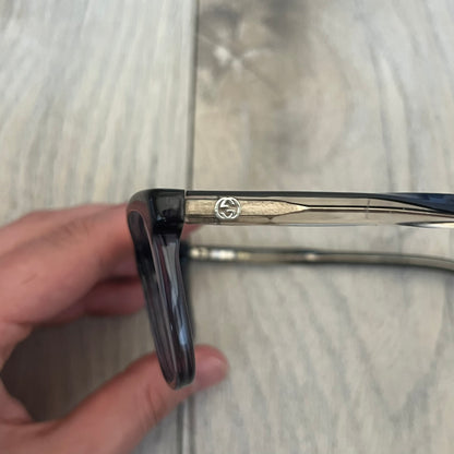 Gucci Gray-Blue Glasses Frames feature a translucent gray and dark blue acetate construction with a clear finish. Small silver GG logos are positioned on the front sides of the frames near the hinges. The frames are sold without lenses and come with a Gucci monogram case and a brown cleaning cloth.