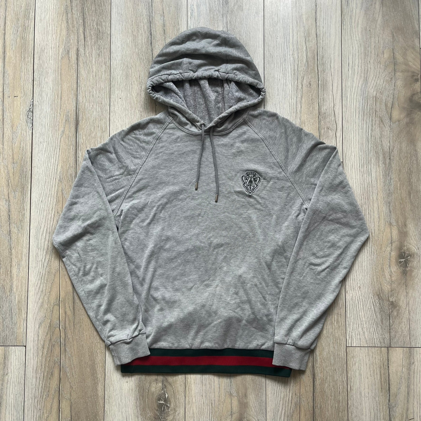 Gucci Gray Crest Patch Sherry Line Zip-Up Hoodie is a lightweight gray zip hoodie made in Italy. The chest features a crest patch with Gucci logo, while the bottom hem includes the green and red Web (Sherry Line) detail. The drawstrings have metal aglets engraved with Gucci branding.