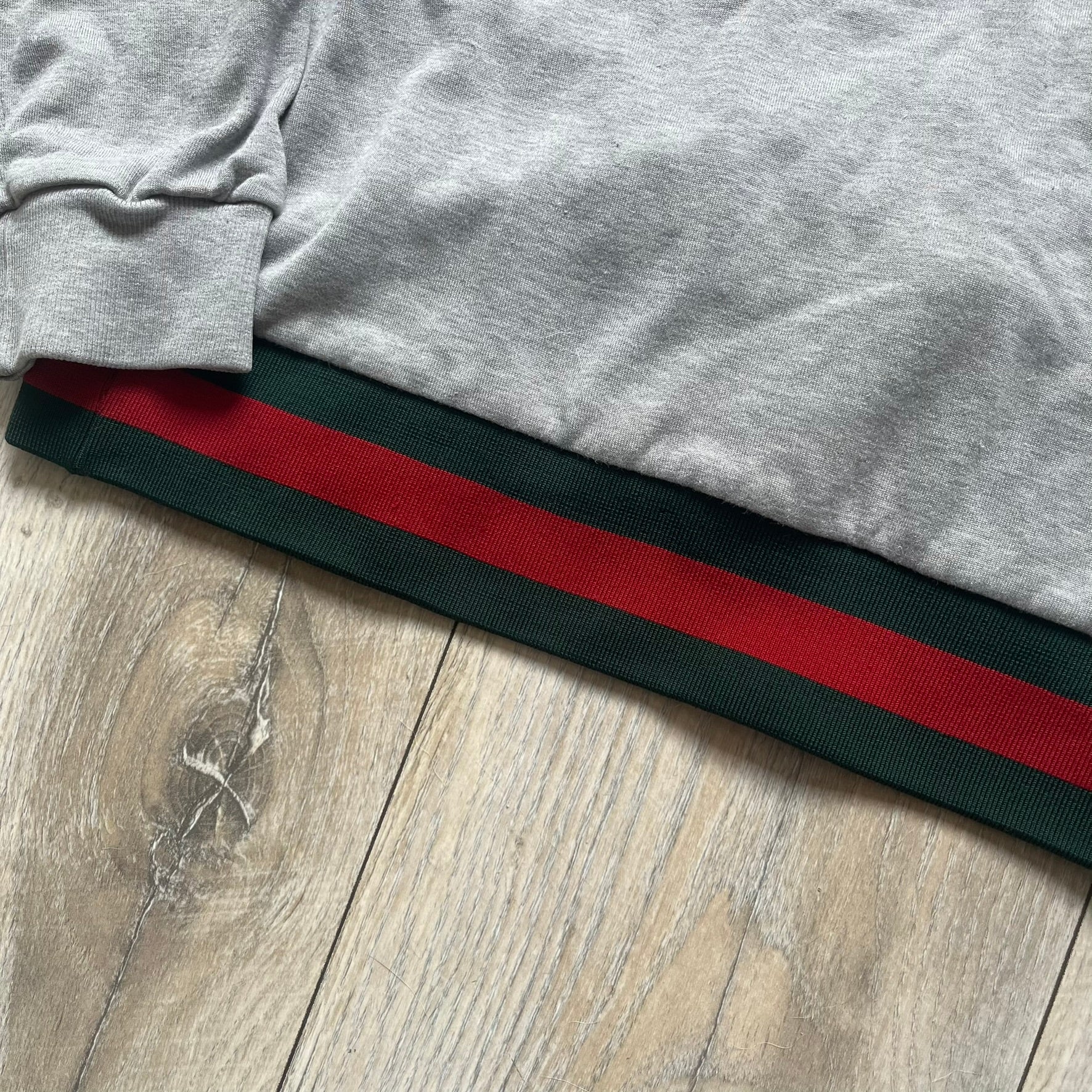 Gucci Gray Crest Patch Sherry Line Zip-Up Hoodie is a lightweight gray zip hoodie made in Italy. The chest features a crest patch with Gucci logo, while the bottom hem includes the green and red Web (Sherry Line) detail. The drawstrings have metal aglets engraved with Gucci branding.