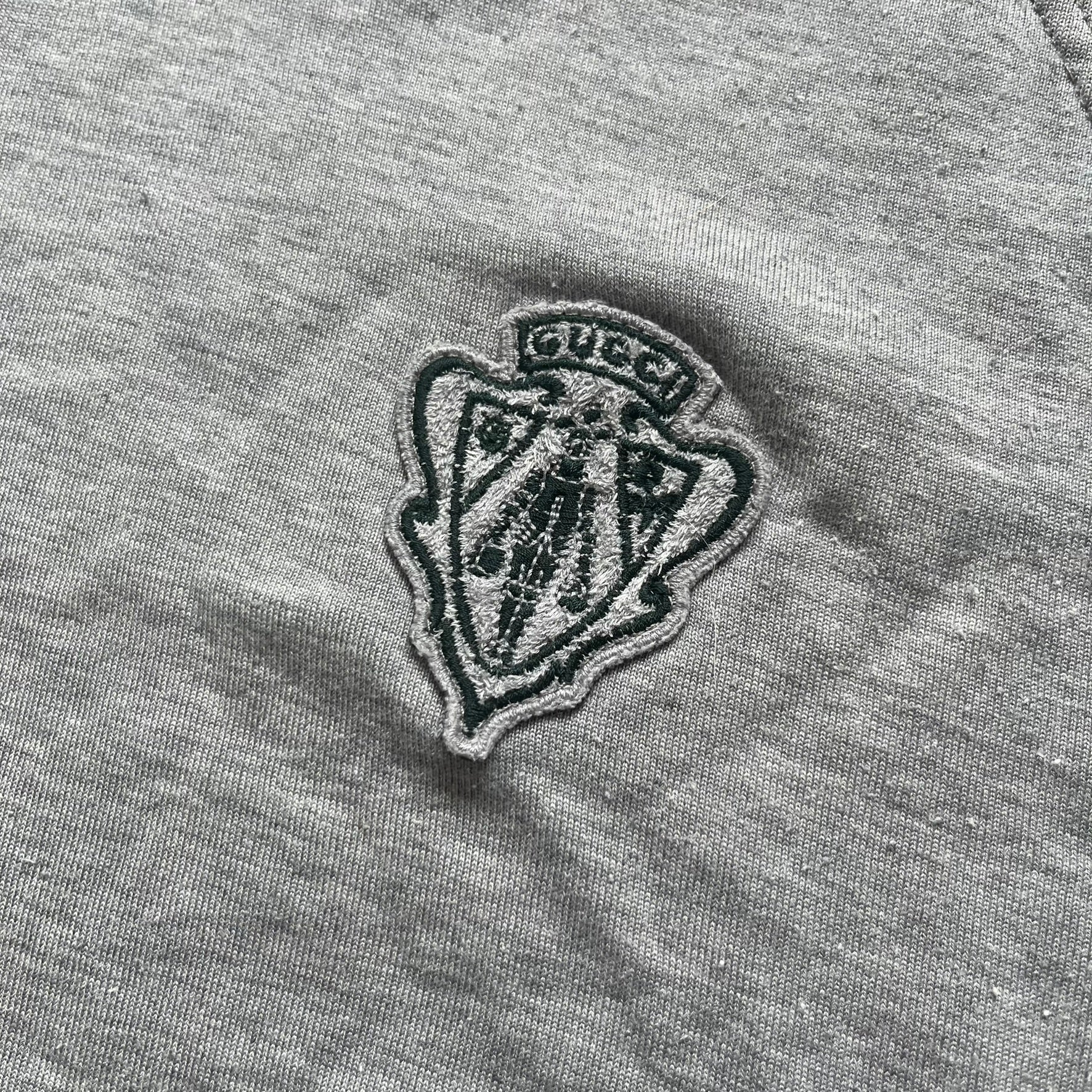 Gucci Gray Crest Patch Sherry Line Zip-Up Hoodie is a lightweight gray zip hoodie made in Italy. The chest features a crest patch with Gucci logo, while the bottom hem includes the green and red Web (Sherry Line) detail. The drawstrings have metal aglets engraved with Gucci branding.