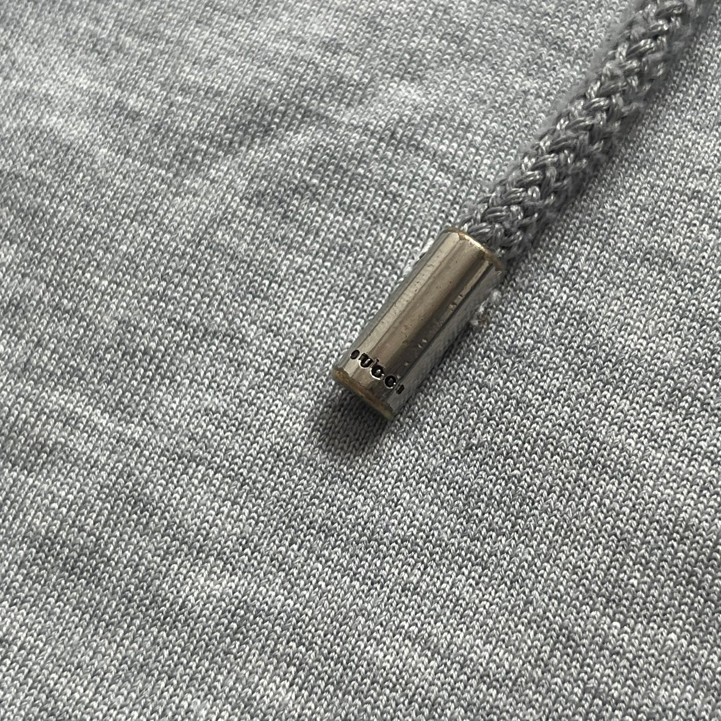 Gucci Gray Crest Patch Sherry Line Zip-Up Hoodie is a lightweight gray zip hoodie made in Italy. The chest features a crest patch with Gucci logo, while the bottom hem includes the green and red Web (Sherry Line) detail. The drawstrings have metal aglets engraved with Gucci branding.
