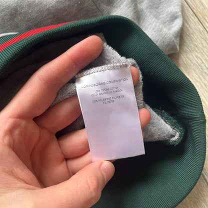 Gucci Gray Crest Patch Sherry Line Zip-Up Hoodie is a lightweight gray zip hoodie made in Italy. The chest features a crest patch with Gucci logo, while the bottom hem includes the green and red Web (Sherry Line) detail. The drawstrings have metal aglets engraved with Gucci branding.