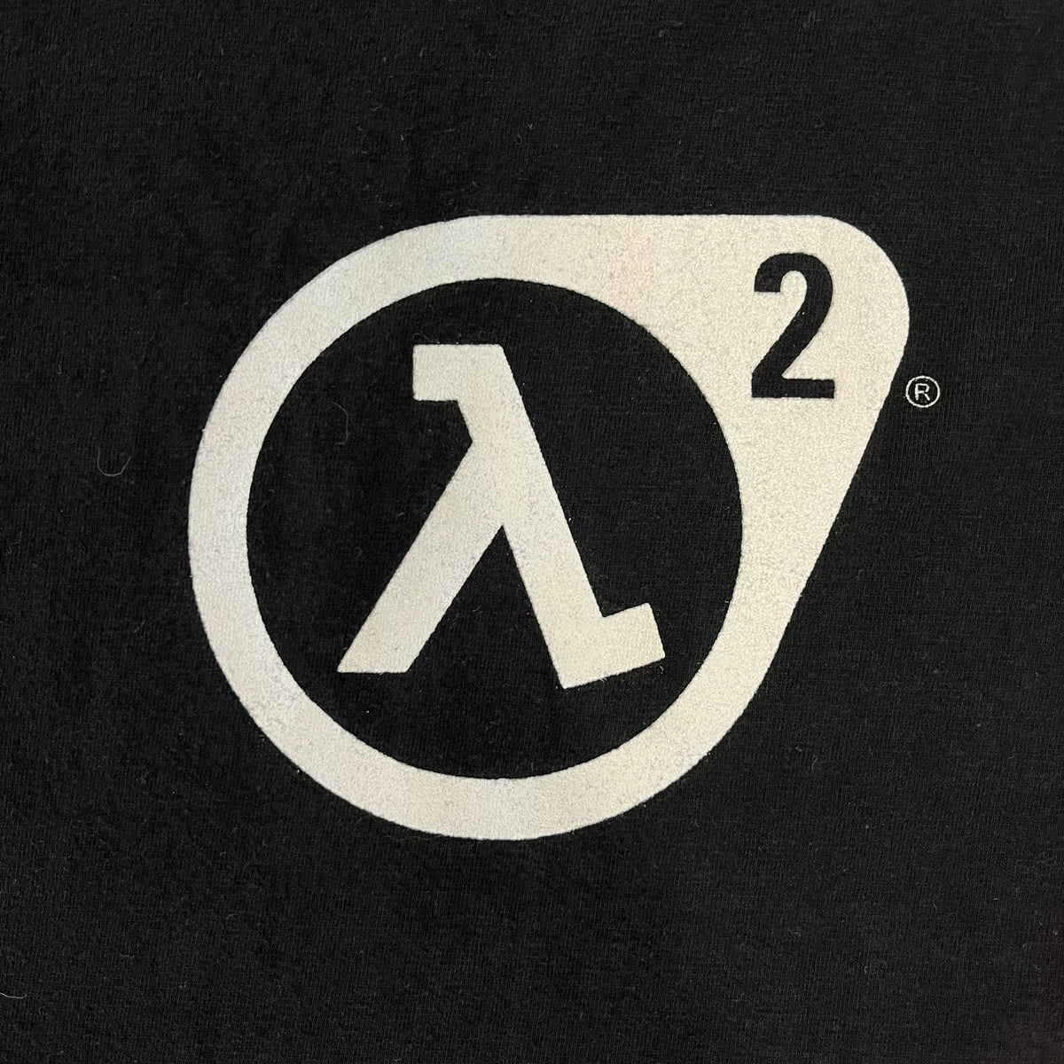 Half-Life 2 2000’s Lambda Black Tee is a black, 100% cotton T-shirt with a white Half-Life 2 logo on the front, showing the lambda symbol inside a circle with a “2” in the corner and a small TM mark beside it.
