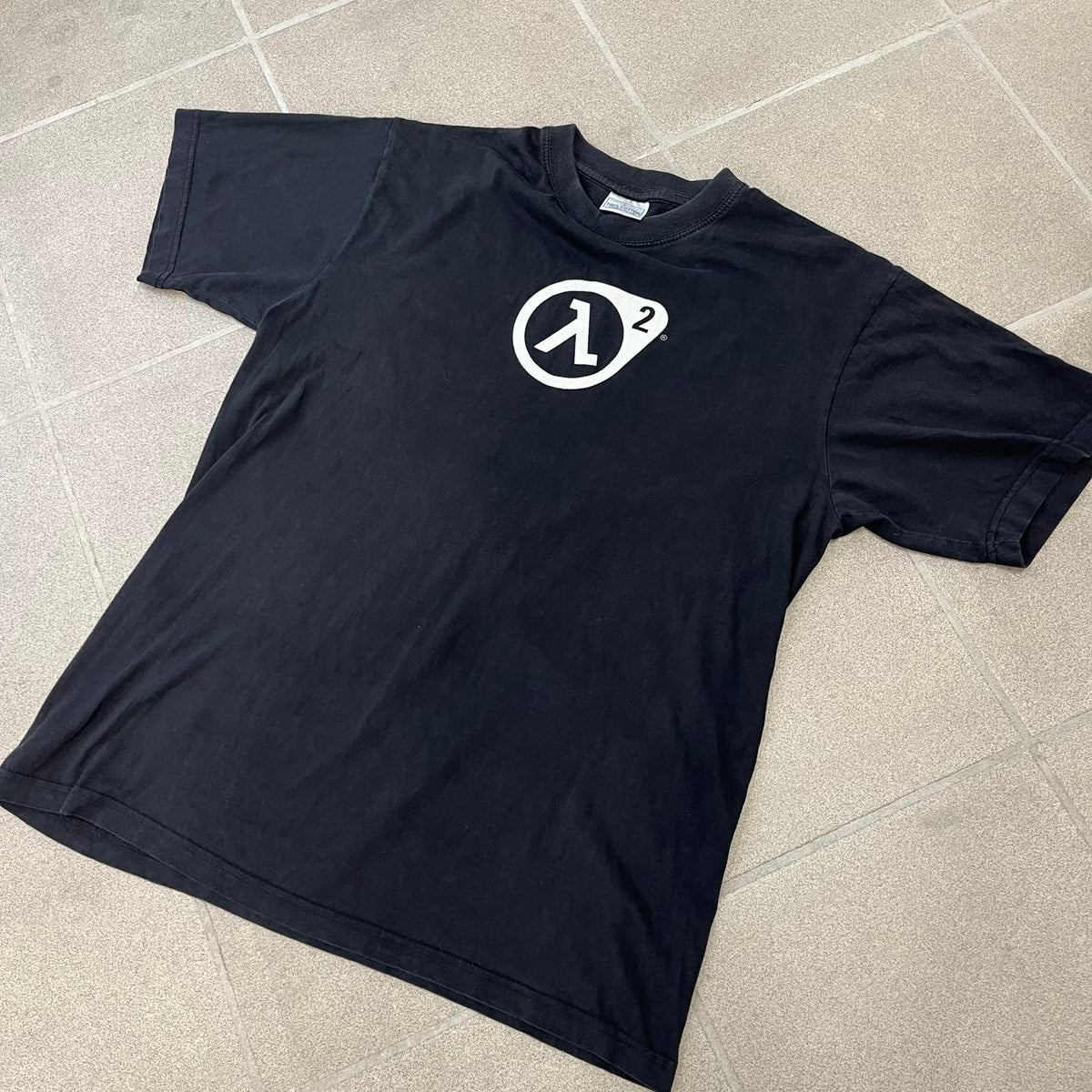 Half-Life 2 2000’s Lambda Black Tee is a black, 100% cotton T-shirt with a white Half-Life 2 logo on the front, showing the lambda symbol inside a circle with a “2” in the corner and a small TM mark beside it.