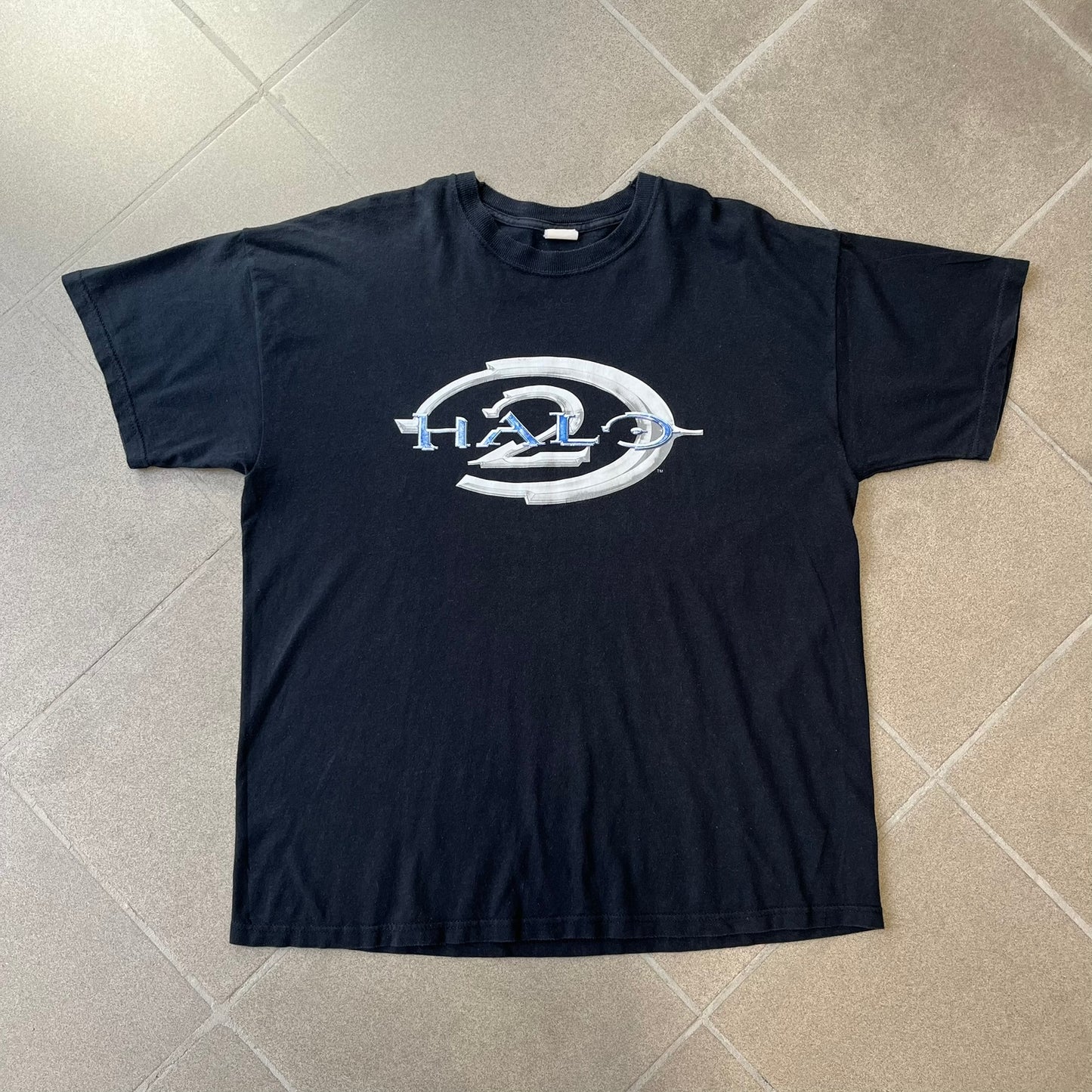 Halo 2 Xbox 2004 Black Tee is a black promotional T-shirt featuring a large white and blue Halo 2 graphic on the front. The upper back has a green original Xbox logo positioned below the collar.