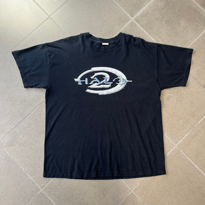 Halo 2 Xbox 2004 Black Tee is a black promotional T-shirt featuring a large white and blue Halo 2 graphic on the front. The upper back has a green original Xbox logo positioned below the collar.