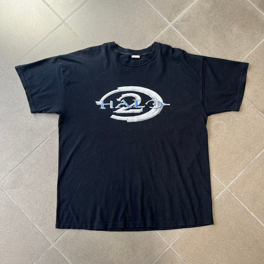 Halo 2 Xbox 2004 Black Tee is a black promotional T-shirt featuring a large white and blue Halo 2 graphic on the front. The upper back has a green original Xbox logo positioned below the collar.